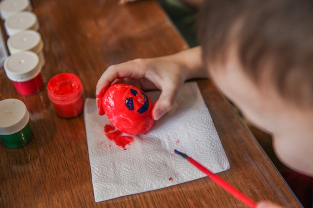 Un bambino dipinge un uovo di pasqua con la tempera rossa e vi disegna una faccia