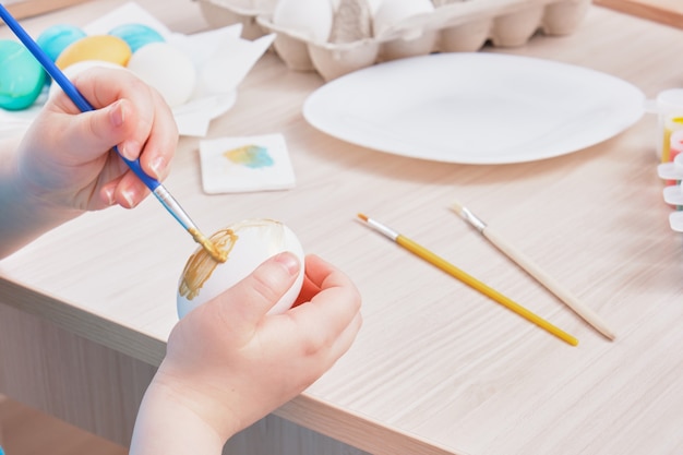 Child paints Easter egg on a wooden table,  eggs box, plate, brushes and paints on the table