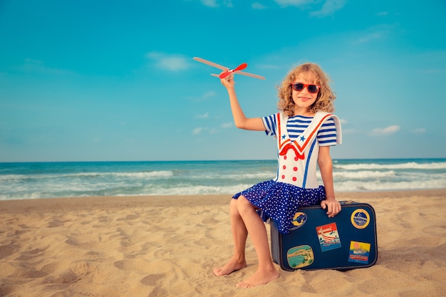 child playing with a airplane and sitting on suitcase at the beach against sea and sky background