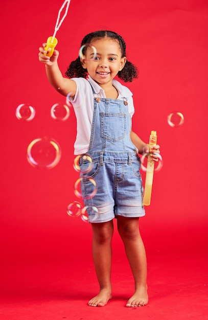 Child portrait or bubbles playing on isolated red background in hand eye coordination kids activity or fun game smile happy or little girl and soap wand studio toy or breathing development skill