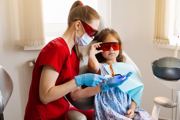 Child prepare for Uv illumination of photopolymer tooth filling in dentistry Dentist in red protective glasses treats and removes caries in kid girl patient