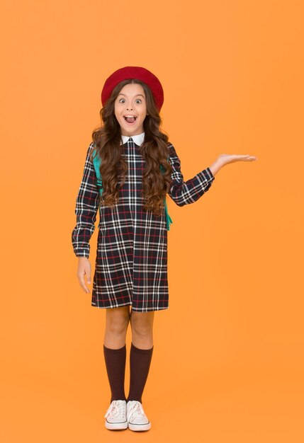 Child in pupil uniform presenting product kid in french beret on yellow background stylish teen college student little girl with backpack going to school education concept school and fashion