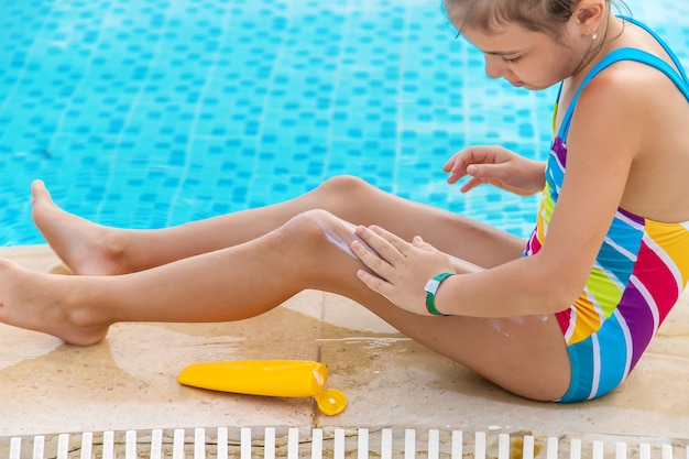 The child puts sunscreen on her leg by the pool