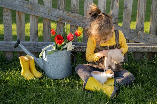 A child in rain boots and country clothes sits by an old wooden fence and a watering can with spring flowers