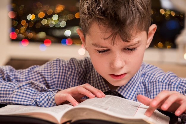 Child reading a thick book. Boy reads book beside window. Young pupil doing homework. Kid solving task in book.