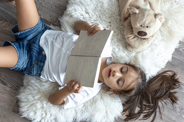 The child reads a book lying on a cozy rug at home with his favorite toy teddy bear.