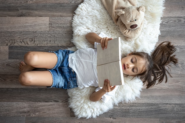 The child reads a book lying on a cozy rug at home with his favorite toy teddy bear.