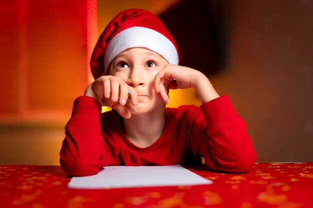 A child in a red jacket on the background of a Christmas tree and garlands writes a letter to Santa Claus