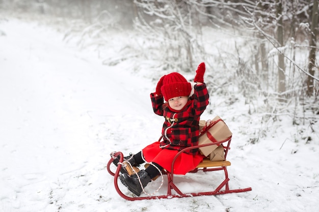 赤い格子縞のコートを着た子供がクリスマスプレゼントを持ってそりに座って笑っています