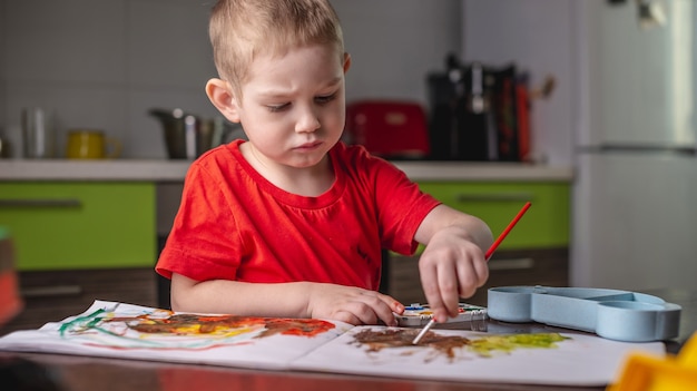 A child in a red T-shirt paints with colorful watercolors at the table