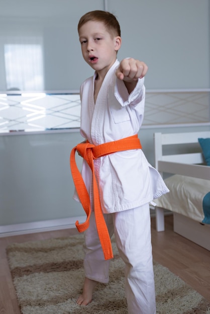 写真 子供の赤毛の少年は、完全に成長しているオレンジ色の帯を持つ着物の形で自宅で空手のトレーニングキックに従事しています子供のスポーツ