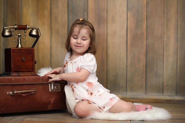 A child in a retro interior and an old phone sits on the floor. A small child a traveler in vintage decorations. Child traveler is calling by phone.