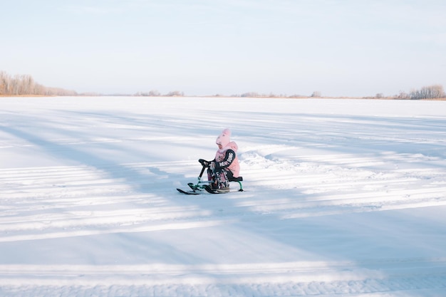 子供は雪道でスノーキャットに乗る ピンクの暖かいジャケットを着た少女は、自然の中を散歩し、晴れた冬の日の側面図に凍った川でそりを楽しんでいます