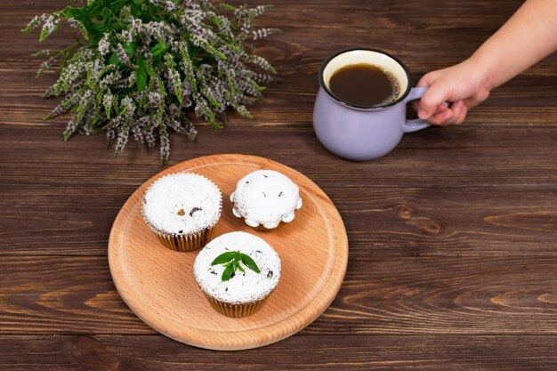 A child's hand holds a cup of tea and mint near the board with muffins with black currant on a background of flowers and mint leaves on a wooden background