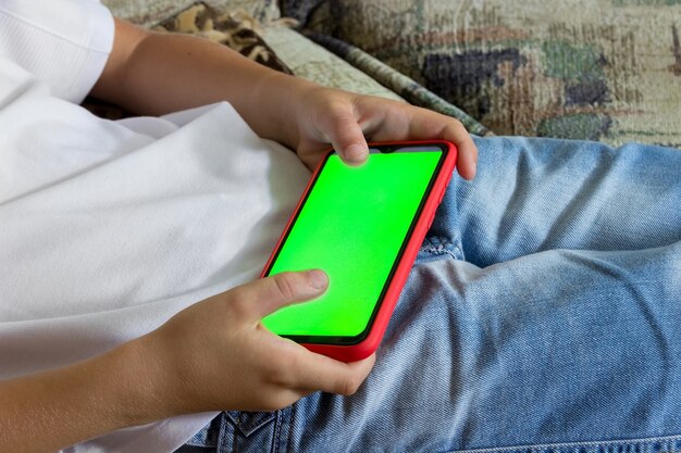 子供の手は、緑色の画面で水平位置にスマートフォンを持っています。クロマキー。モックアップ