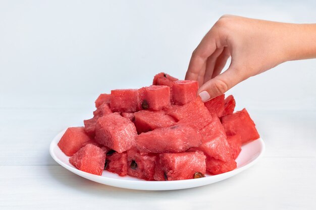 Child's hand takes juicy pieces of ripe red watermelon from a white plate on the table.