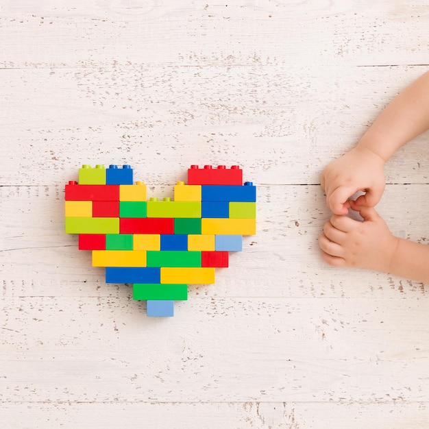 Детские руки играют с ярким сердцем из разноцветных пластиковых кирпичей на старом деревянном столе