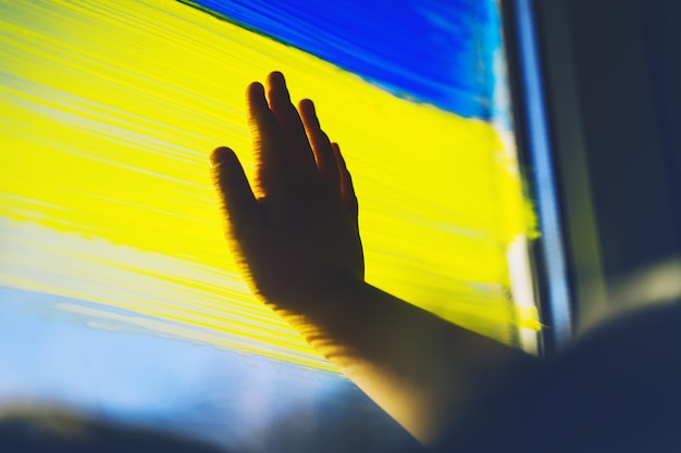 아이의 손이 창에 우크라이나의 노란색 파란색 깃발을 만집니다. 우크라이나 개념을 지원합니다.