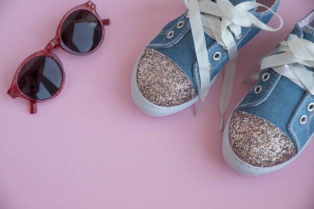 Child's textile lace sneakers. girls shoes on pink wall.Fashion kid's footwear.Smart casual fashionable denim and shiny shoes.Sunglasses and trendy kids sports shoes.Copy space. Selective focus