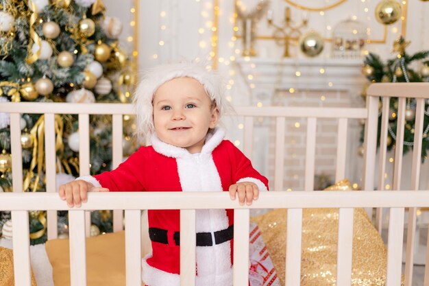 A child in a Santa costume stands in a crib at home near a Christmas tree with a Golden decor