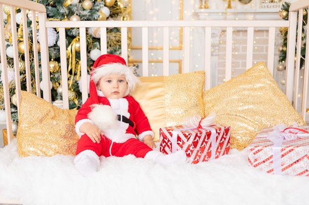 A child in a Santa costume with a gift at the Christmas tree with a Golden decor is sitting in a crib at home