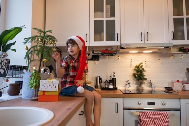 A child in a Santa hat is sitting in a festive kitchen decorated for Christmas A happy girl dresses up a houseplant araucaria as a Christmas tree New Year