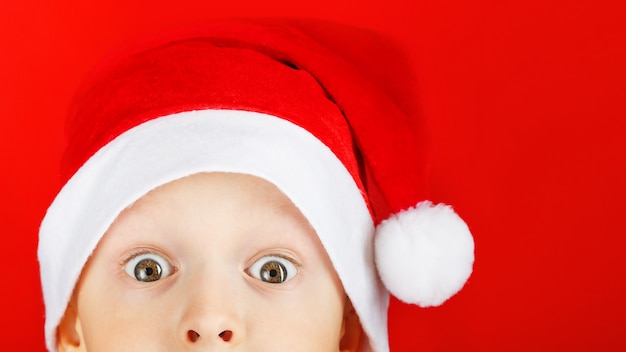 Child in a Santa hat on a red background looks out in surprise from the bottom of the frame