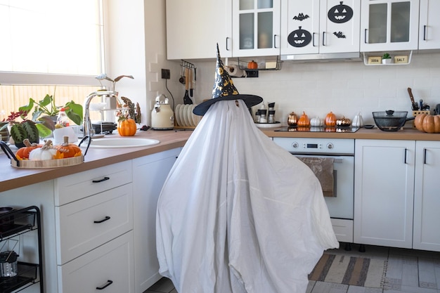 A child in sheets with cutout for eyes like a ghost costume in the kitchen decorated for the Halloween holiday A kind little funny ghost Halloween Party