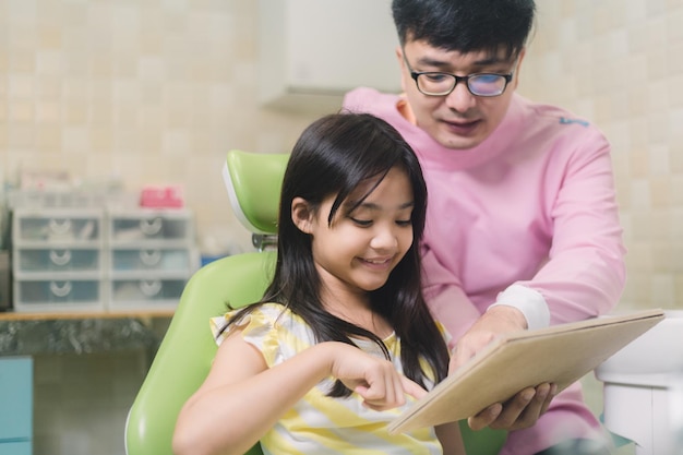 A child sits in a dentist chair and reads a book to a young girl.