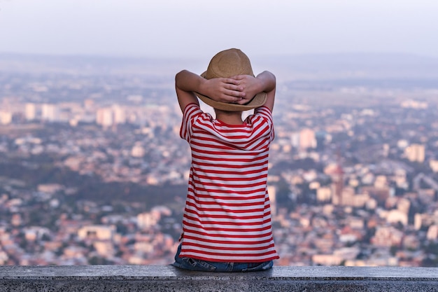 Child sits in panorama city background and his hands behind head Back view