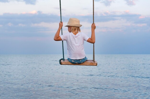 Child sits on swing above the water and looks out to sea Happy childhood The rest of the sea