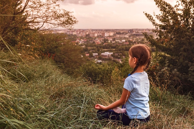 The child sits in a yoga pose on a hill on a background of cityscape