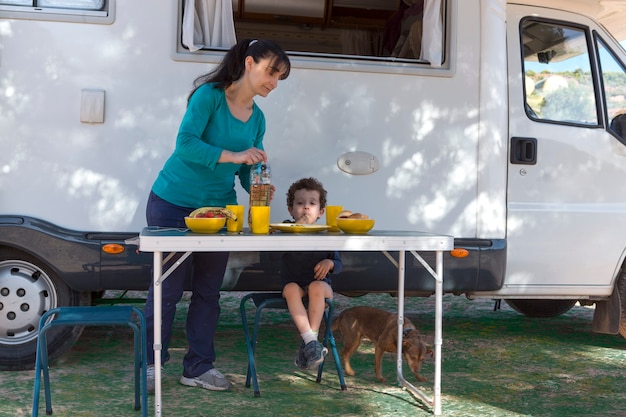 Child sitting at the table next to the camper, while his mother serves snacks and drinks on a camping day.