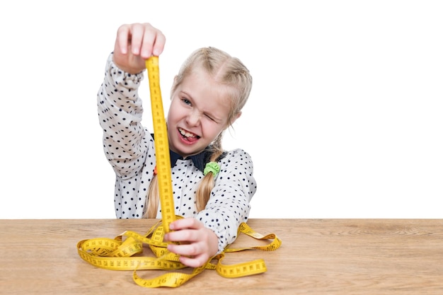 Child sitting at a wooden table measures using a yellow tape measure isolated on a white background