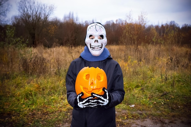 Foto bambino in maschera teschio e guanti scheletrici che tengono jack'o'lantern di zucca di halloween