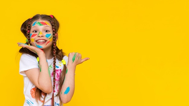 A child smeared with paint a little girl painted with multicolored paints points to your advertisement children's creativity yellow isolated background copy space banner