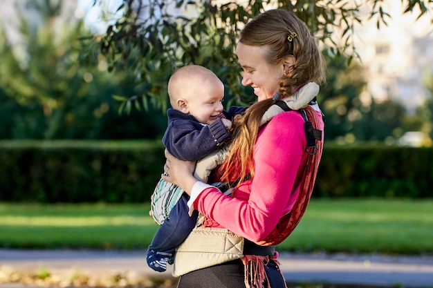 Child smiles in baby sling in hands of his mother during walking in park