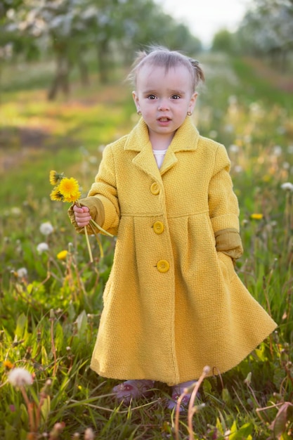 Bambino nel giardino di primavera bambina divertente in un cappotto giallo sta tenendo i denti di leone