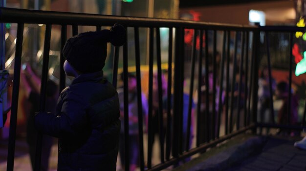 A child standing alone holding on to the fence looking for something