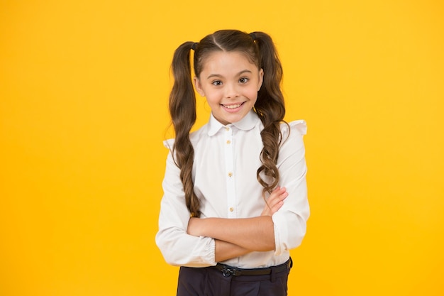 Child starting school Happy small child keeping arms crossed on yellow background Little child smiling with long brunette hair in formal school uniform Cheerful female child back to school