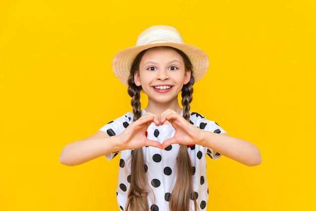 A child in a straw hat shows a heart with his fingers A beautiful little girl in a polka dot Tshirt is smiling broadly while enjoying the summer Yellow isolated background