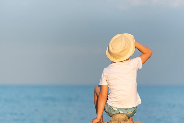 Child in a straw hat sitting on beach and looking at the distance