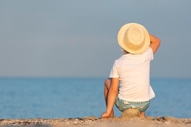 Child in a straw hat sitting on beach and looking at sea