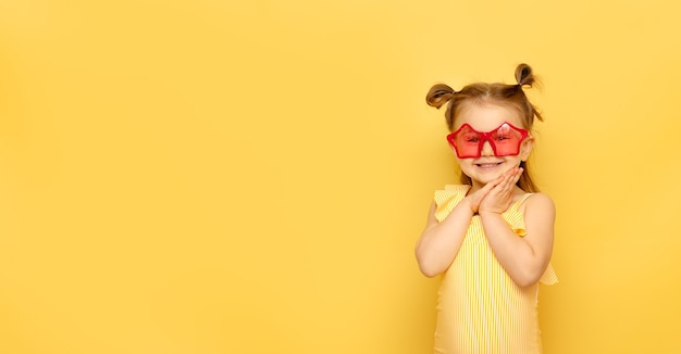 Child in striped swimsuit and red funny summer sunglasses looks at camera posing on yellow wall