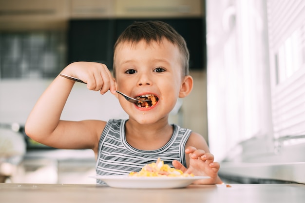 Un bambino in maglietta in cucina che mangia una frittata con salsiccia e pomodori con una forchetta