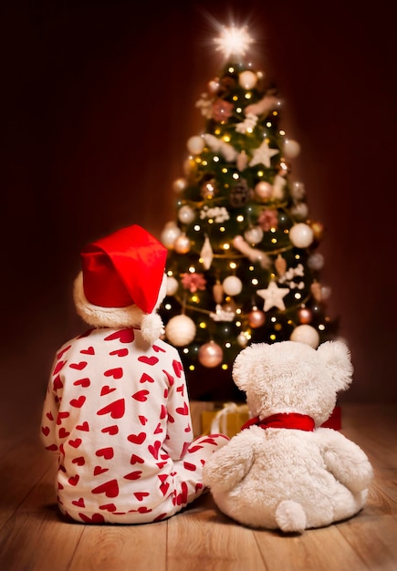 a child and a teddy bear sit on the background of the Christmas tree