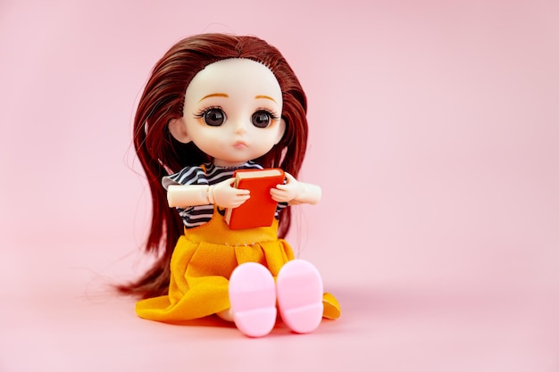 A child toy doll with big eyes with dark hair in an orange dress sitting on a pink background Plastic children's toy Doll games for imagination copy space