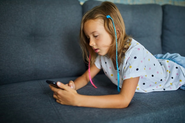 Photo child using cellphone on the sofa in modern and bright apartment, young girl plays on smartphone at home, problem of gadget addiction