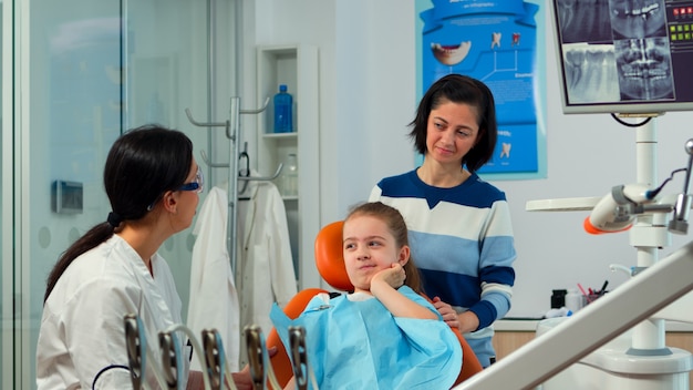 Bambino che usa il dito per puntare il dente affetto mentre il dentista parla con la madre del mal di denti orale. medico di odontoiatria che spiega alla mamma il processo dentale, figlia seduta su una sedia stomatologica