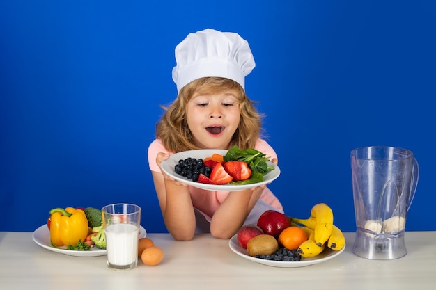 Child wearing cooker uniform and chef hat hold plate with fruits preparing vegetables on kitchen stu